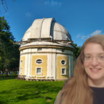 a photograph of an old observatory dome building in white and yellow with green grass around it and a blue sky, to the right a blonde woman with long hair and glasses