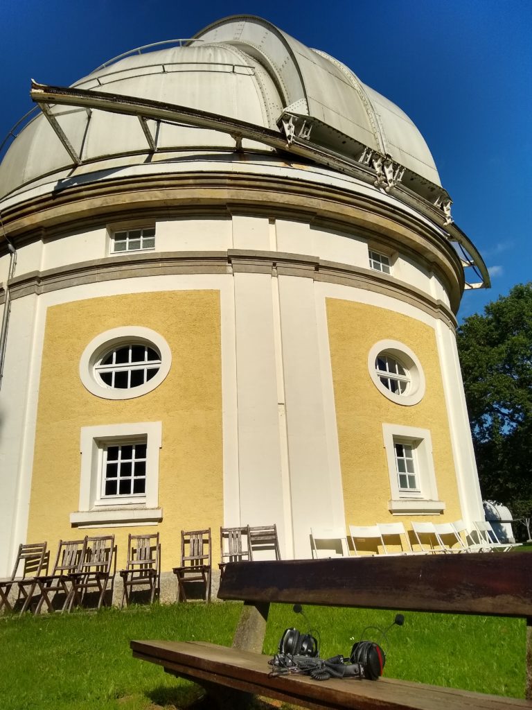 Photograph of an old white and yellow observatory dome in the sun with blue sky around and a bench surrounded by green grass in the front with podcast recording equipment on it