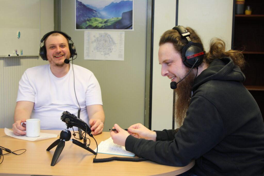 Two young men with headsets and microphones are sitiing at a desk and smiling
