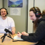 Two young men with headsets and microphones are sitiing at a desk and smiling