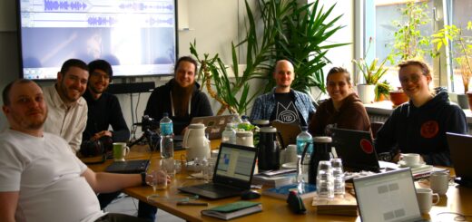 Seven smiling people sitting around a table with laptops, coffee flasks and water bottles, looking into the camera. Screen with audio editing software mounted on the wall behind them.