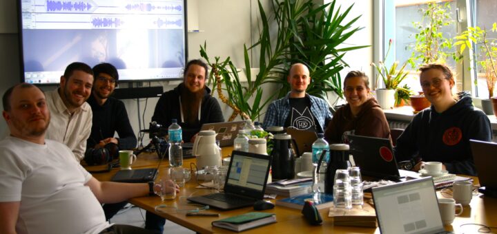 Seven smiling people sitting around a table with laptops, coffee flasks and water bottles, looking into the camera. Screen with audio editing software mounted on the wall behind them.