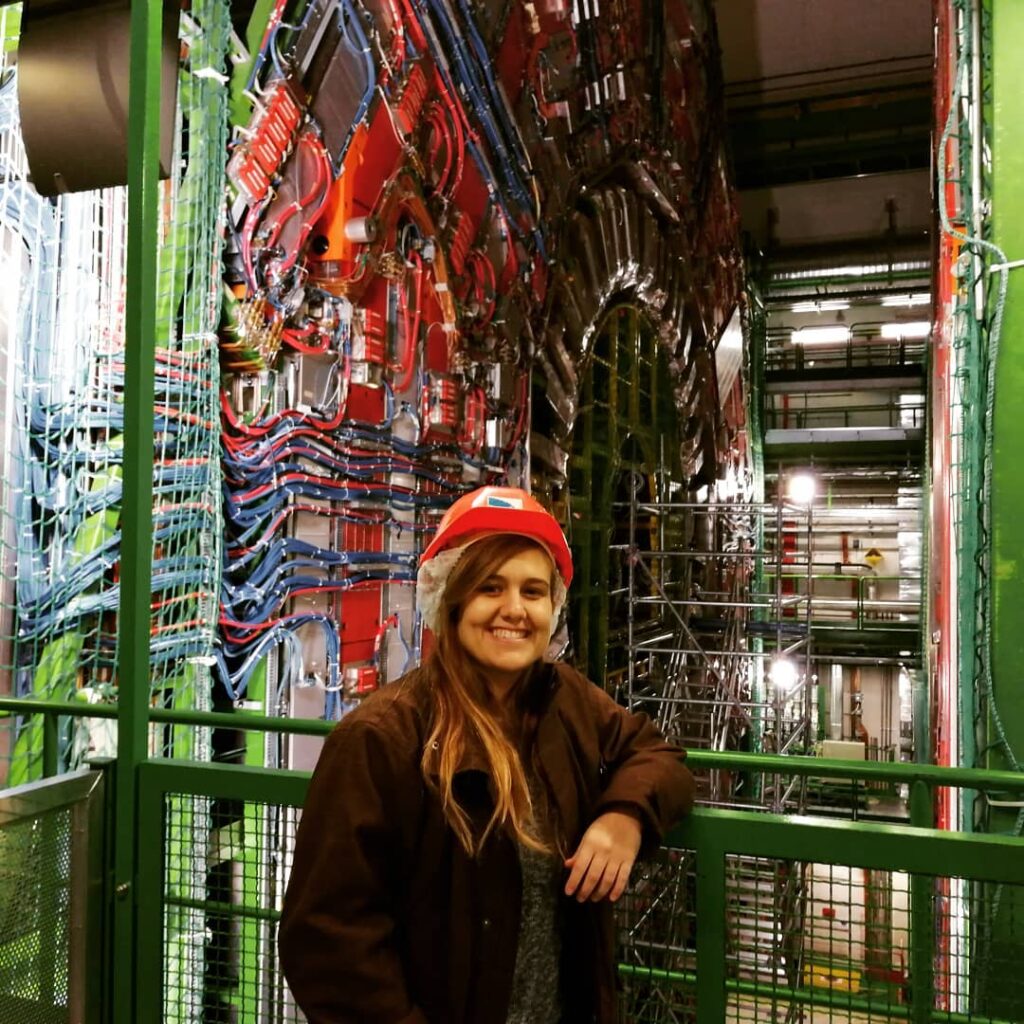 photo of a smiling young woman wearing a helmet and standing in front of a huge particle detector