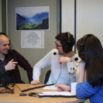 Two men and one woman sitting at a table wearing headphones, engaged in lively conversation